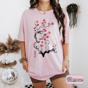 May include: A light pink t-shirt featuring a black ink-style illustration of a tree branch with bright pink cherry blossoms and three small white forest spirit figures. The design includes black Japanese kanji characters and a small red circular stamp.