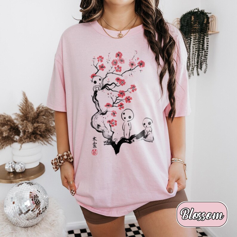 May include: A light pink t-shirt featuring a black ink-style illustration of a tree branch with bright pink cherry blossoms and three small white forest spirit figures. The design includes black Japanese kanji characters and a small red circular stamp.