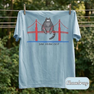 May include: Light blue t-shirt featuring a cartoon cat sitting on the Golden Gate Bridge in San Francisco. The bridge is red, and the text "SAN FRANCISCO" is below the bridge. The shirt is hanging on a clothesline.
