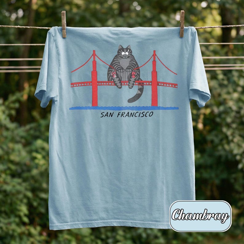 May include: Light blue t-shirt featuring a cartoon cat sitting on the Golden Gate Bridge in San Francisco. The bridge is red, and the text "SAN FRANCISCO" is below the bridge. The shirt is hanging on a clothesline.