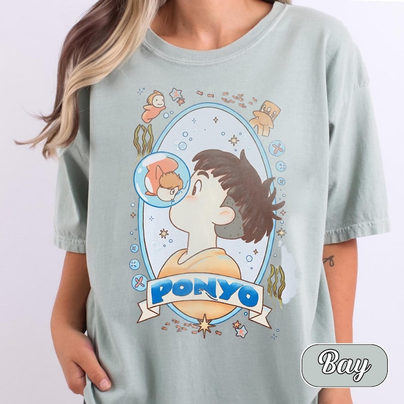 May include: A sage green short-sleeve t-shirt featuring a graphic illustration of a character with brown hair and an orange shirt, kissing a small orange fish inside a bubble. The word PONYO is printed in blue letters on a white banner below.