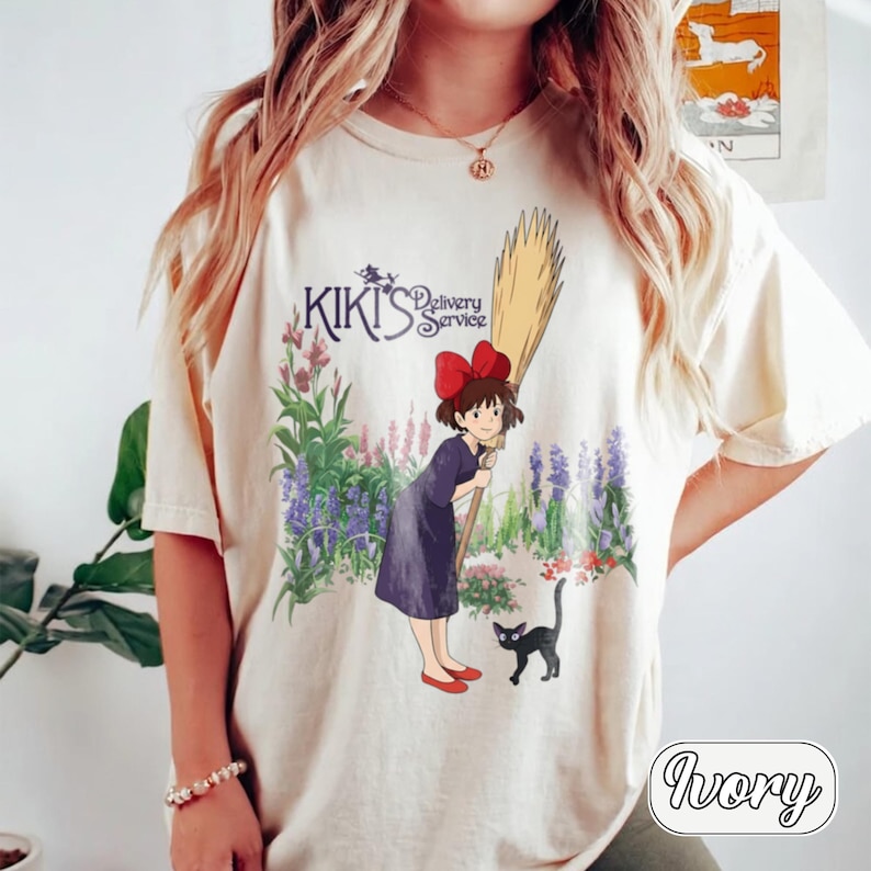 May include: An ivory short-sleeve t-shirt featuring a graphic of Kiki from Kiki's Delivery Service holding a broomstick, accompanied by a black cat. The design includes purple and green flowers and the text Kiki's Delivery Service in dark blue.