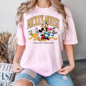 May include: A light pink t-shirt featuring a graphic of Mickey Mouse and friends. The text "MICKEY MOUSE" is arched across the top, with "FRIENDS FOREVER" below. The cartoon characters are in bright colors.