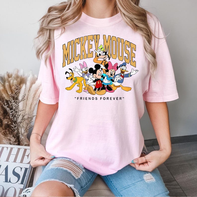 May include: A light pink t-shirt featuring a graphic of Mickey Mouse and friends. The text "MICKEY MOUSE" is arched across the top, with "FRIENDS FOREVER" below. The cartoon characters are in bright colors.
