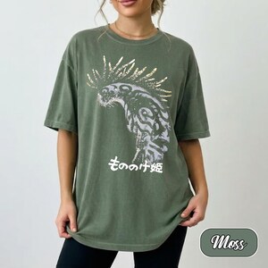 May include: A moss green short-sleeve t-shirt featuring a graphic print of a stylised creature with a spiky mane in shades of grey and white. Below the creature, the Japanese text "もののけ姫" is printed in white. The shirt has a crew neckline.