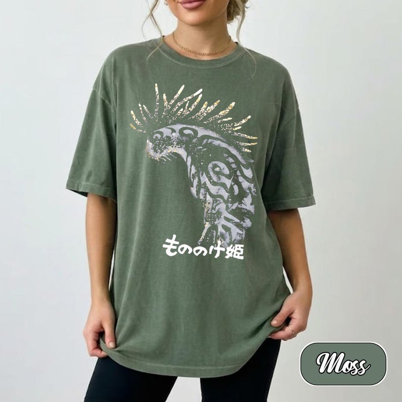 May include: A moss green short-sleeve t-shirt featuring a graphic print of a stylised creature with a spiky mane in shades of grey and white. Below the creature, the Japanese text "もののけ姫" is printed in white. The shirt has a crew neckline.