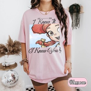 May include: A light pink short-sleeve t-shirt featuring a graphic print of a cartoon character with red hair and a blue sky background. The text reads EST 2026, I Knew It, and I Knew You with a musical note symbol at the end of the final line.
