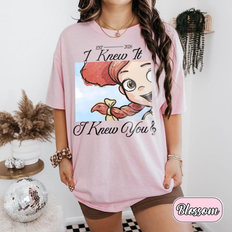 May include: A light pink short-sleeve t-shirt featuring a graphic print of a cartoon character with red hair and a blue sky background. The text reads EST 2026, I Knew It, and I Knew You with a musical note symbol at the end of the final line.