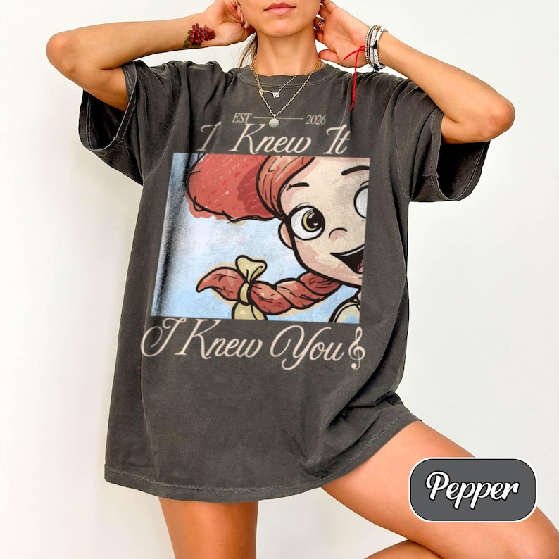 May include: A charcoal gray short-sleeve t-shirt featuring a graphic print of a cartoon character with red hair in braids. The text reads "EST 2026", "I Knew It", and "I Knew You" with a musical note symbol. The shirt has a relaxed, loose fit.