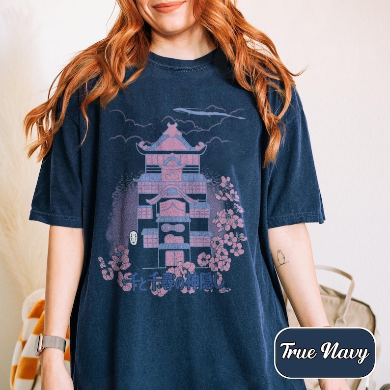 May include: A navy blue short-sleeve t-shirt featuring a graphic print of a multi-story building with pink and blue accents, surrounded by cherry blossoms and clouds. The Japanese text "千と千尋の神隠し" is printed below the building illustration.