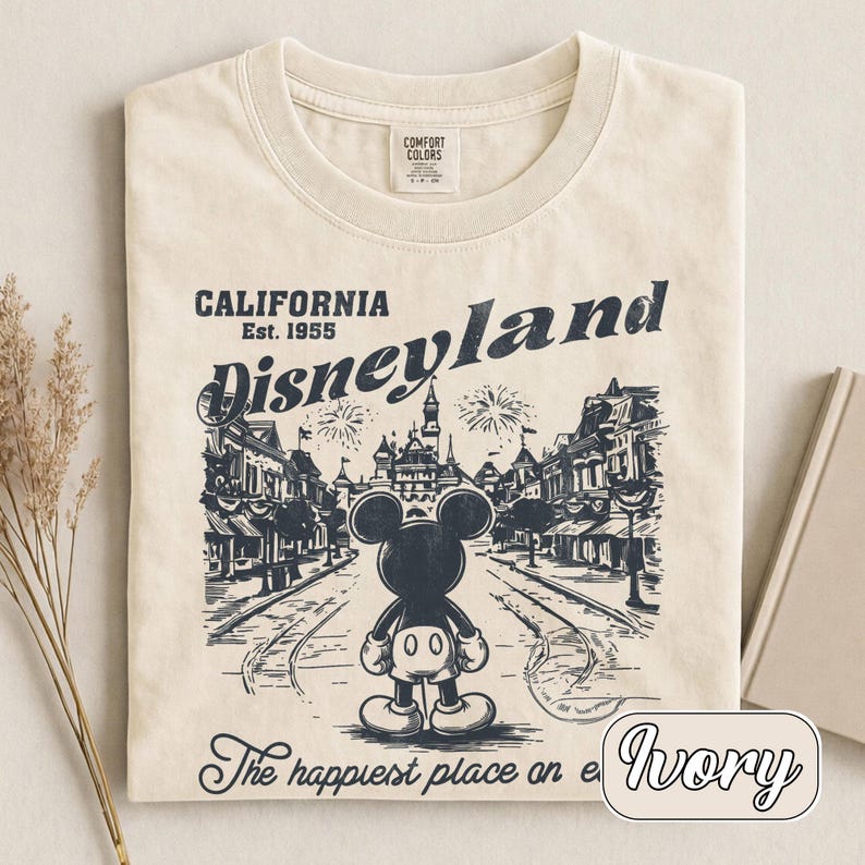 May include: Ivory-colored t-shirt featuring a Disneyland graphic. The design includes Mickey Mouse, the Disneyland logo, and the text "California Est. 1955." The phrase "The happiest place on earth" is also printed on the shirt.