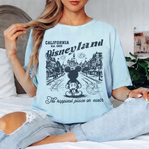 May include: Light blue t-shirt with a Disneyland graphic. The graphic features Mickey Mouse standing in front of a street scene with the text "California Est. 1955 Disneyland" and "The happiest place on earth."