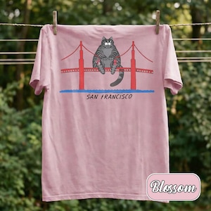 May include: A light pink t-shirt featuring a cartoon cat sitting on the Golden Gate Bridge in San Francisco. The bridge is red, and the text "SAN FRANCISCO" is below the bridge. The shirt is hanging on a clothesline.