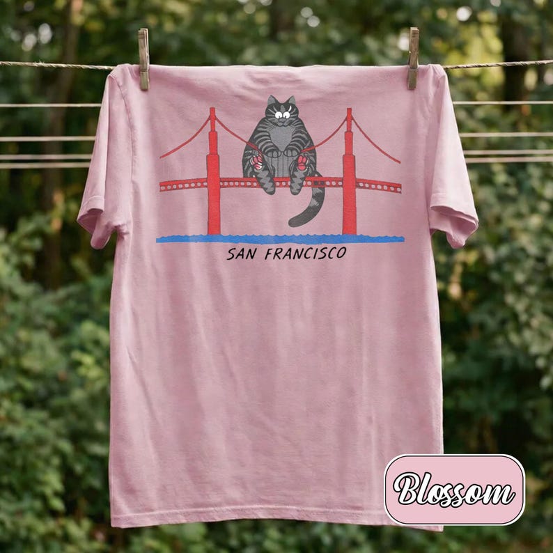 May include: A light pink t-shirt featuring a cartoon cat sitting on the Golden Gate Bridge in San Francisco. The bridge is red, and the text "SAN FRANCISCO" is below the bridge. The shirt is hanging on a clothesline.