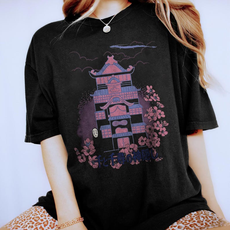 May include: A black short-sleeve t-shirt featuring a graphic print of a multi-story Japanese-style building in shades of pink and purple. The design includes cherry blossom branches, a small white mask figure, and Japanese text at the bottom.