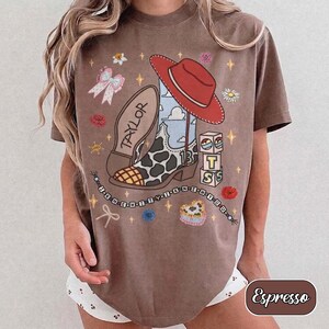 Pu&ograve; includere: Una t-shirt marrone a maniche corte con una stampa grafica raffigurante uno stivale da cowboy maculato, un cappello rosso, un fiocco e blocchi con le lettere T, S e il numero 13. Il testo TAYLOR &egrave; presente sullo stivale e perline.