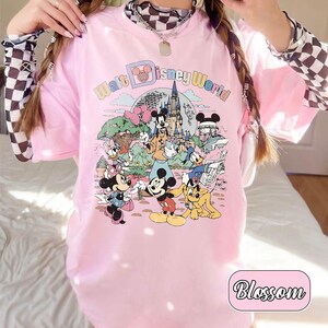 May include: Pink t-shirt featuring a colorful graphic of Disney characters and the text "Walt Disney World." The design includes Mickey Mouse, Minnie Mouse, Donald Duck, and other characters in a cartoon style. The word "Blossom" is in a pink rounded rectangle.