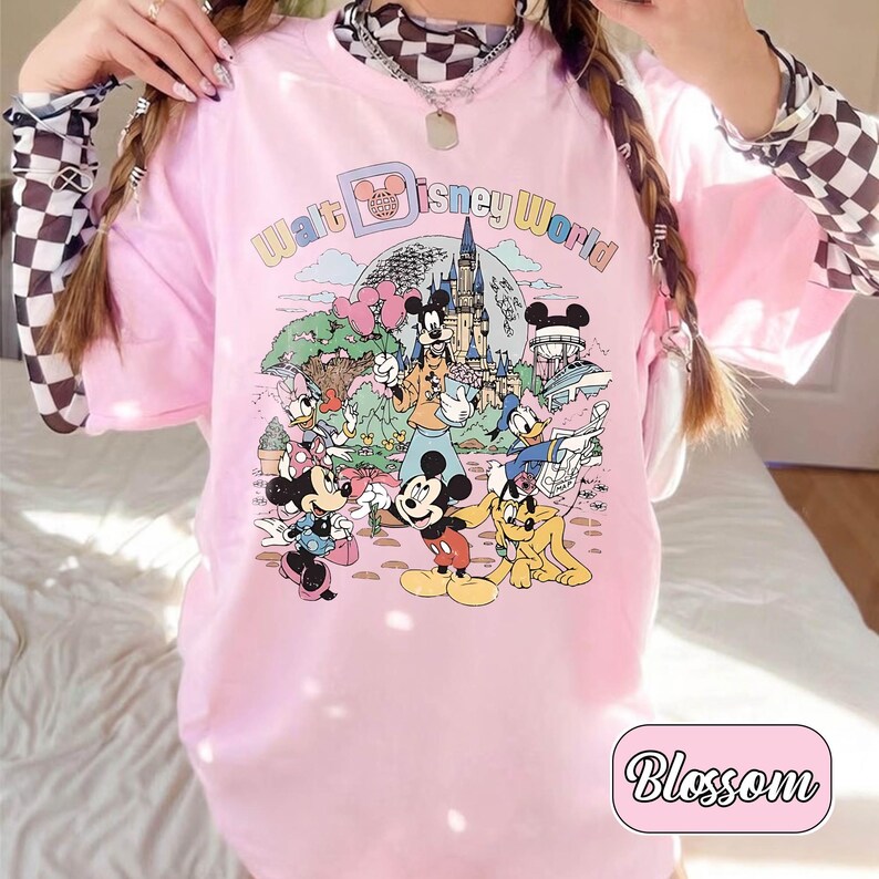 May include: Pink t-shirt featuring a colorful graphic of Disney characters and the text "Walt Disney World." The design includes Mickey Mouse, Minnie Mouse, Donald Duck, and other characters in a cartoon style. The word "Blossom" is in a pink rounded rectangle.