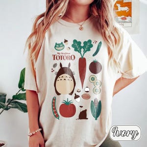 May include: An ivory short-sleeve t-shirt featuring a graphic print of My Neighbor Totoro characters and various vegetables. The design includes a brown Totoro, a red tomato, a green carrot, corn, and other garden produce in a colorful illustration.
