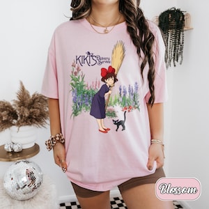 May include: A light pink t-shirt featuring a graphic print of Kiki from Kiki's Delivery Service, holding a broomstick with a black cat nearby, set against a floral background. The text Kiki's Delivery Service is printed in purple above the character.