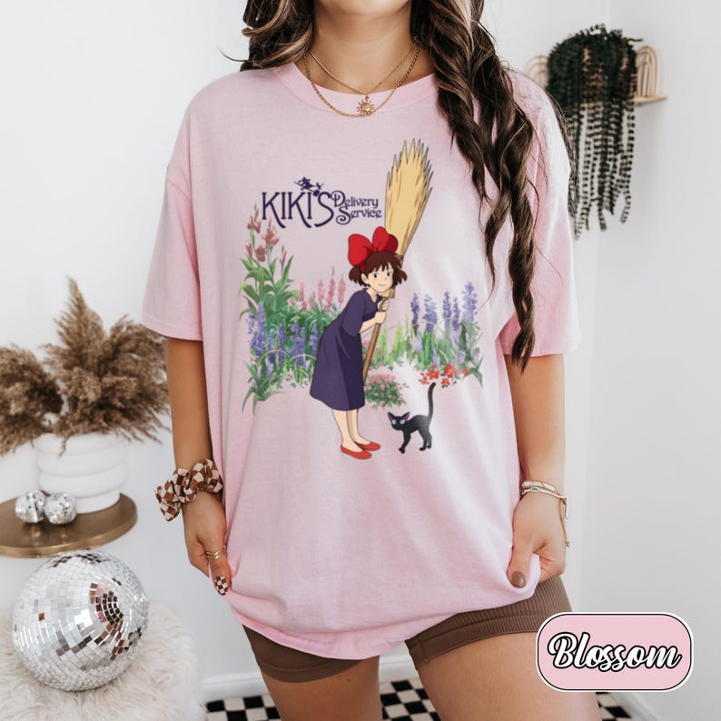 May include: A light pink t-shirt featuring a graphic print of Kiki from Kiki's Delivery Service, holding a broomstick with a black cat nearby, set against a floral background. The text Kiki's Delivery Service is printed in purple above the character.