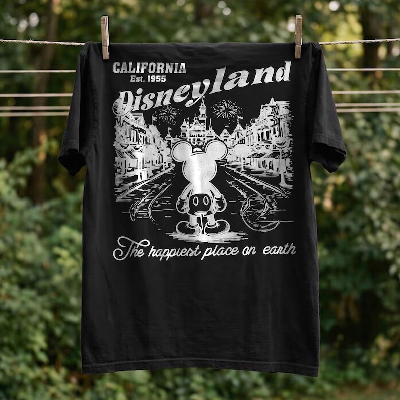 May include: Black t-shirt featuring a Disneyland graphic. The design includes the words "California Est. 1955" and "Disneyland" above an illustration of Mickey Mouse and a street scene. The text "The happiest place on earth" is below.
