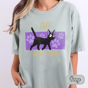May include: A light sage green t-shirt featuring a graphic of a black cat named Jiji. The design includes a purple rectangular background with light purple paw prints, the text "JIJI" in yellow, and Japanese characters in yellow below the cat.
