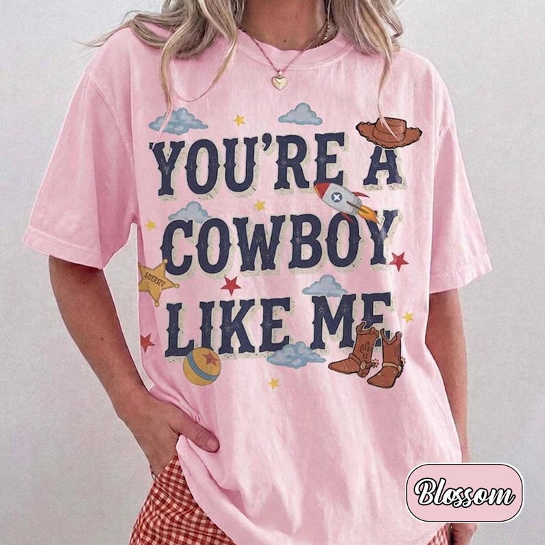 K&ouml;nnte beinhalten: Ein hellrosa T-Shirt mit kurzen &Auml;rmeln und dem dunkelblauen Schriftzug "YOU'RE A COWBOY LIKE ME", umgeben von grafischen Illustrationen eines braunen Cowboyhuts, einer Rakete, eines gelben Sheriffsterns, eines bunten Balls und braunen Cowboystiefeln.