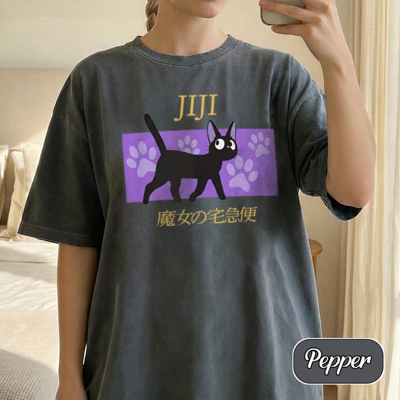 May include: A charcoal gray short-sleeve t-shirt featuring a graphic of a black cat named Jiji. The design includes a purple rectangular background with white paw prints, the text Jiji, and Japanese characters below the cat illustration.
