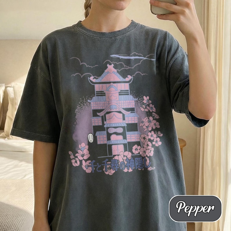 May include: A charcoal gray t-shirt featuring a graphic print of a multi-story building in shades of pink and blue, surrounded by cherry blossoms and clouds. The Japanese text "千と千尋の神隠し" is printed below the building, with a small white mask icon.