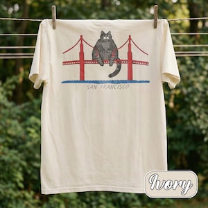 May include: Ivory-colored t-shirt featuring a gray cat sitting on the Golden Gate Bridge, with red towers and the text "SAN FRANCISCO" below. The shirt is hanging on a clothesline.