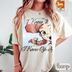 May include: An ivory t-shirt featuring a graphic print of a cartoon character with red hair and a yellow bow. The text reads "EST 2026 I Knew It I Knew You" with a musical note symbol. The design is centered on the chest of the short-sleeved garment.
