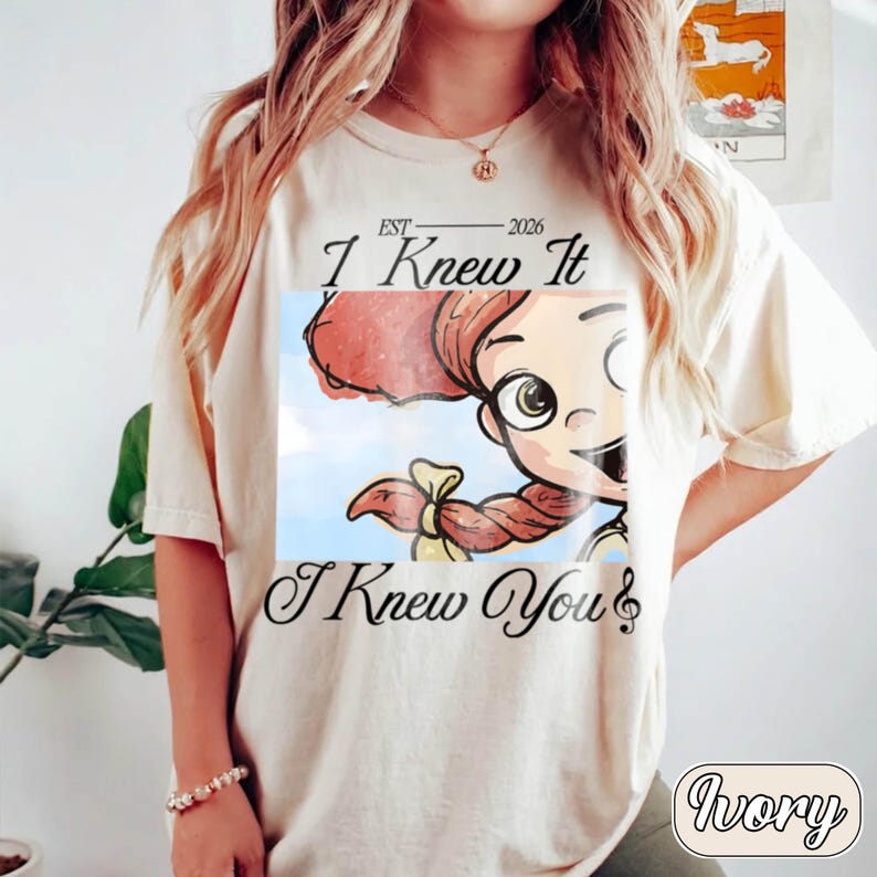May include: An ivory t-shirt featuring a graphic print of a cartoon character with red hair and a yellow bow. The text reads "EST 2026 I Knew It I Knew You" with a musical note symbol. The design is centered on the chest of the short-sleeved garment.