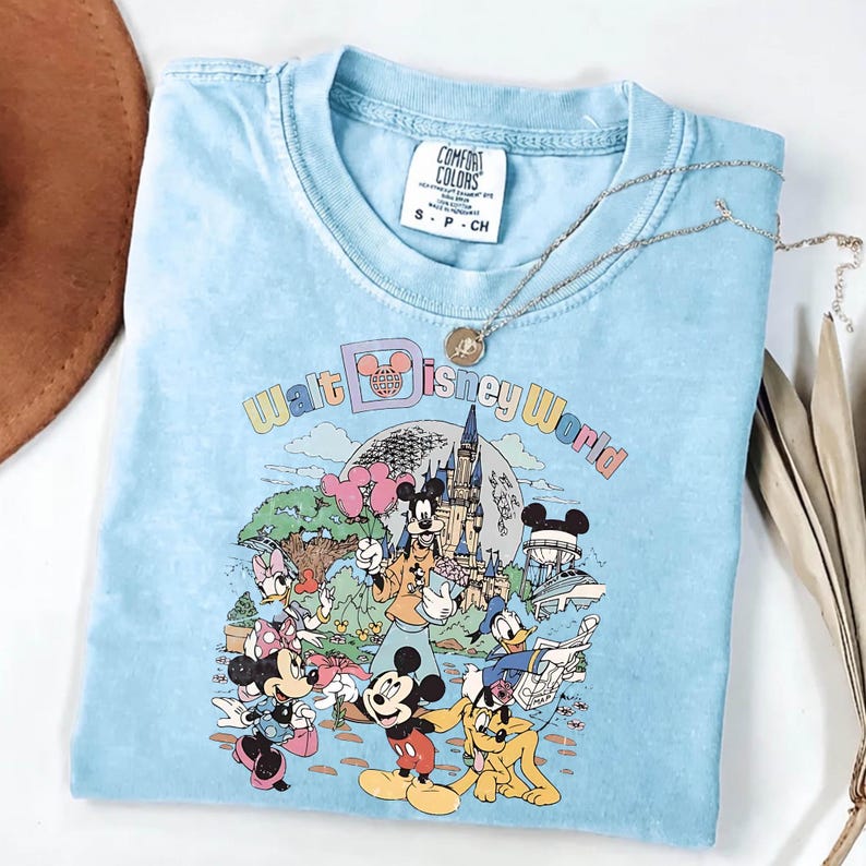 May include: Light blue t-shirt featuring a colorful graphic of Disney characters and the text "Walt Disney World." The shirt has a crew neck and short sleeves. A silver necklace with a pendant is draped across the shirt.