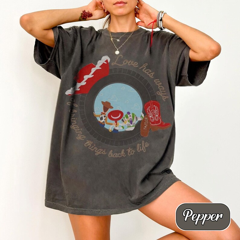 May include: A charcoal gray short-sleeve t-shirt featuring a circular graphic with cartoon characters, a red cowboy boot, and the text "Love has ways of bringing things back to life." The shirt has a loose fit and a crew neckline.