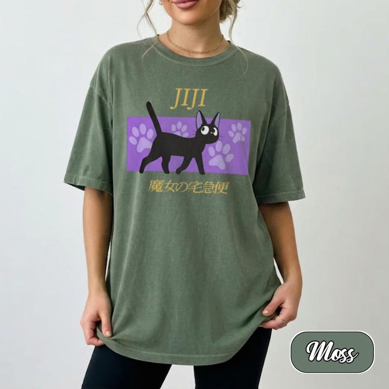 May include: A moss green short-sleeve t-shirt featuring a graphic of a black cat walking across a purple rectangular background with paw prints. The text JIJI is printed above the cat, and Japanese characters are printed below the cat.