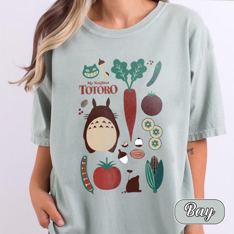 May include: A light sage green t-shirt featuring a graphic print of the character Totoro from My Neighbor Totoro, surrounded by various vegetables including a carrot, tomato, corn, and peas, along with acorns and small forest elements.