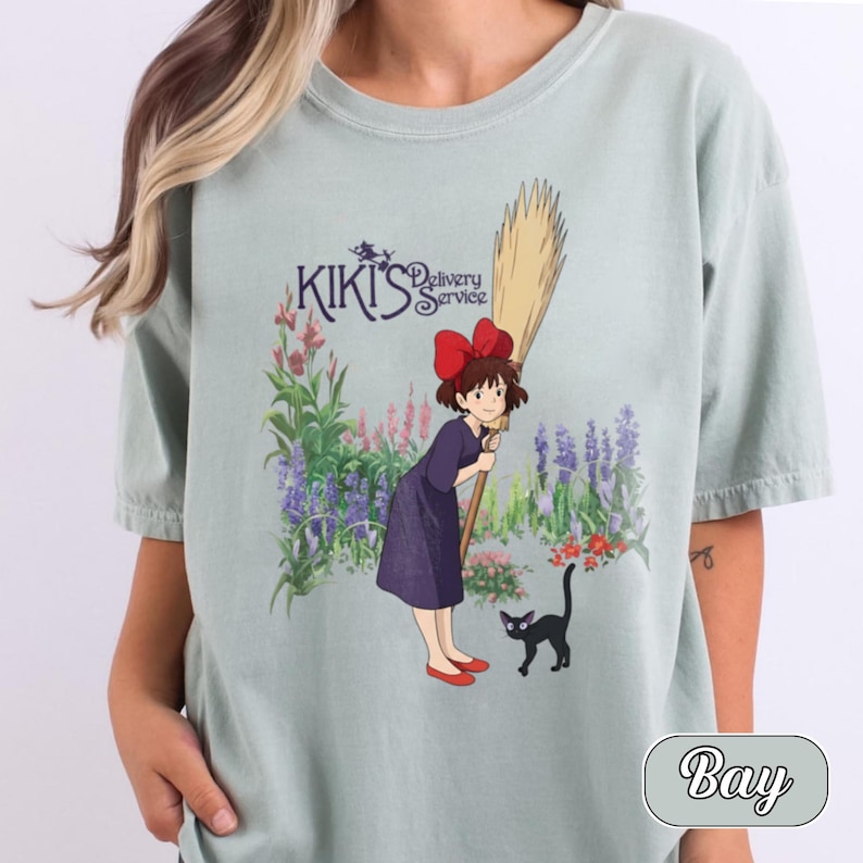 May include: A light sage green t-shirt featuring a graphic print of Kiki from Kiki's Delivery Service. The design shows Kiki in a dark purple dress with a red bow, holding a broomstick, standing next to a black cat in a garden with flowers.