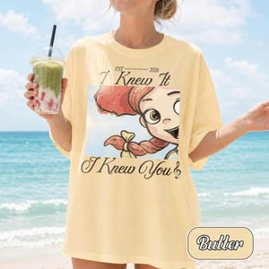 May include: A pale yellow t-shirt featuring a graphic print of a cartoon character with red hair and a braid. The text reads EST 2026 I Knew It I Knew You. The shirt has a crew neckline and short sleeves, presented in a casual, loose-fitting style.