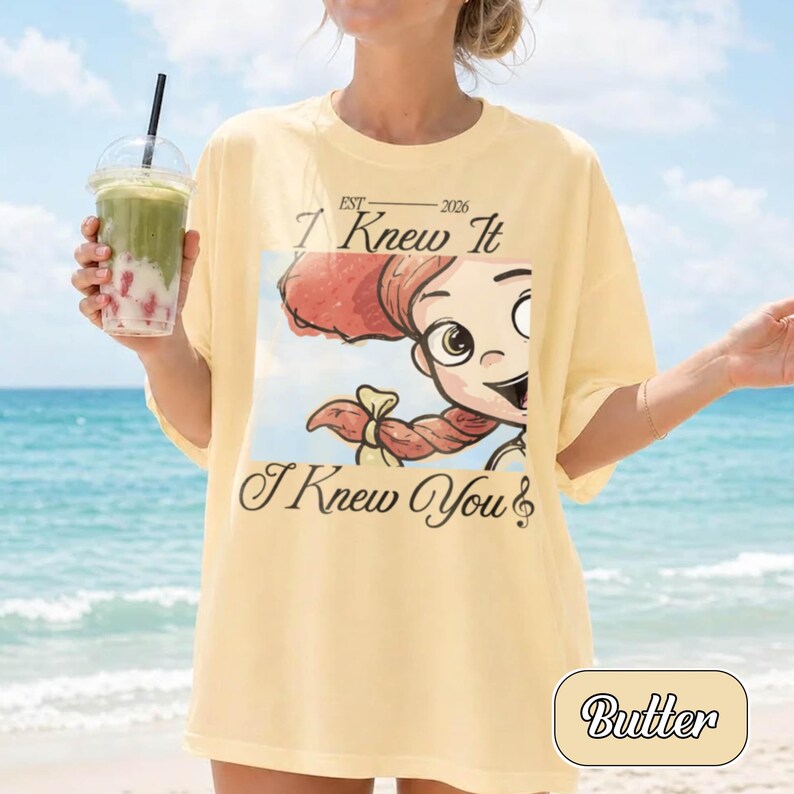 May include: A pale yellow t-shirt featuring a graphic print of a cartoon character with red hair and a braid. The text reads EST 2026 I Knew It I Knew You. The shirt has a crew neckline and short sleeves, presented in a casual, loose-fitting style.