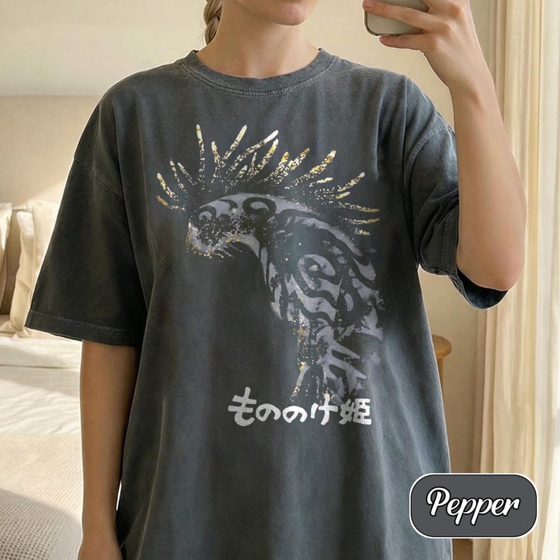May include: A charcoal grey short-sleeve t-shirt featuring a graphic print of a stylised creature with gold-toned accents and the Japanese text "もののけ姫" printed in white below the design. The shirt has a crew neckline and a relaxed fit.