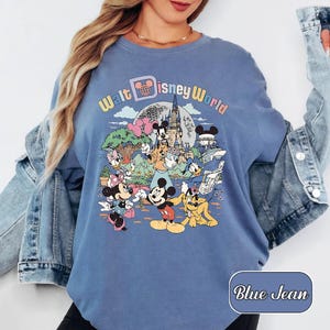 May include: A blue crew neck t-shirt featuring a colorful graphic of Disney characters and the text "Walt Disney World". The design includes Mickey Mouse, Minnie Mouse, Donald Duck, and Goofy. The shirt has a relaxed fit.