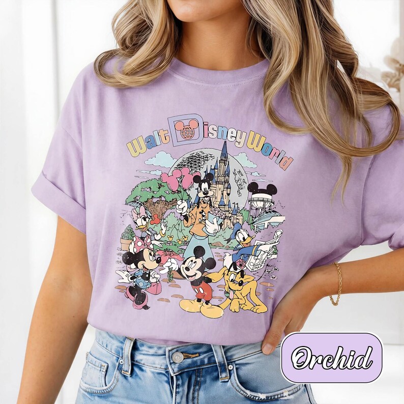 May include: A lilac-colored t-shirt featuring a colorful graphic of Disney characters, including Mickey Mouse, Minnie Mouse, Donald Duck, and Goofy, in front of a castle. The text "Walt Disney World" is arched above the scene.