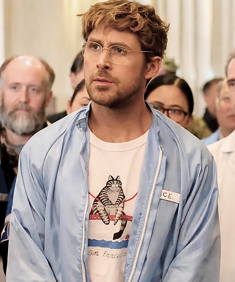May include: A light blue zip-up jacket with a white t-shirt featuring a striped cat graphic and the text "Sm Francisco". The person is wearing round, gold-rimmed glasses.