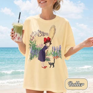May include: A pale yellow t-shirt featuring a graphic print of a character with a red bow holding a broom, a black cat, and purple and green flowers. The text Kiki's Delivery Service is printed above the character in a dark purple font.