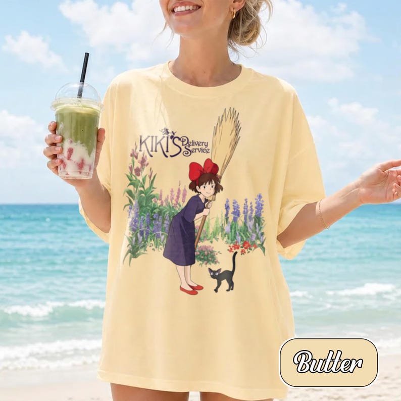 May include: A pale yellow t-shirt featuring a graphic print of a character with a red bow holding a broom, a black cat, and purple and green flowers. The text Kiki's Delivery Service is printed above the character in a dark purple font.