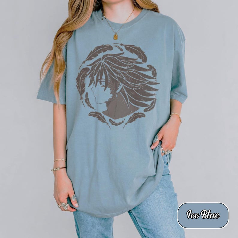Pu&ograve; includere: Una t-shirt a maniche corte color blu ghiaccio con una stampa grafica marrone scuro di un personaggio in stile anime di profilo, circondato da una disposizione circolare di piume. La maglietta ha un girocollo e una vestibilit&agrave; ampia.