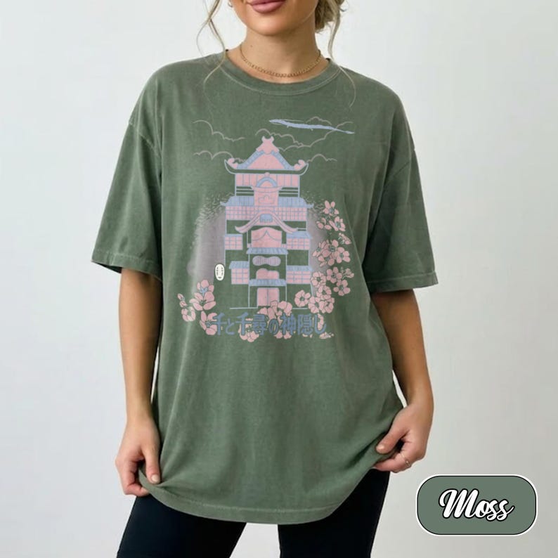 May include: A moss green short-sleeve t-shirt featuring a graphic print of a multi-story building in shades of pink and blue, surrounded by cherry blossom branches. The design includes Japanese text and a small white mask icon near the base.