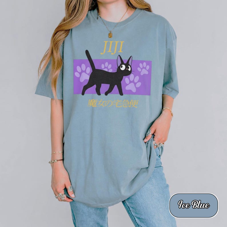 May include: An ice blue t-shirt featuring a graphic of a black cat walking across a purple rectangular background with light purple paw prints. The text JIJI is printed in yellow above the cat, and Japanese characters are printed below.