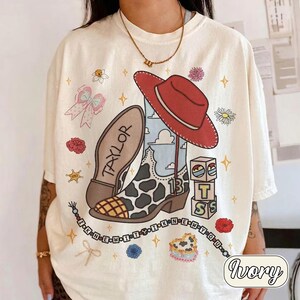 Pu&ograve; includere: Una t-shirt color avorio con una stampa grafica di uno stivale da cowboy con motivo a macchie di mucca, un cappello rosso, blocchi con le lettere T, S e il numero 13, un fiocco e una fila di perline con il testo I KNEW IT Y I KNEW YOU.